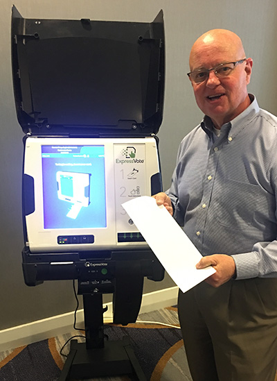 Johnson KS Metsker_ExpressVote White man with bald head and glasses wears a blue collard shirt and holds a blank white ballot card next to an ExpressVote voting machine with a blue and white screen that displays an image of the machine. The voting machine is white and sitting atop a black rolling cart with a black hood